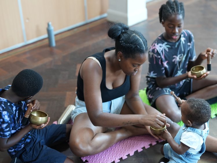 mother with three children sitting on wooden floor each playing tibetan singing bowls