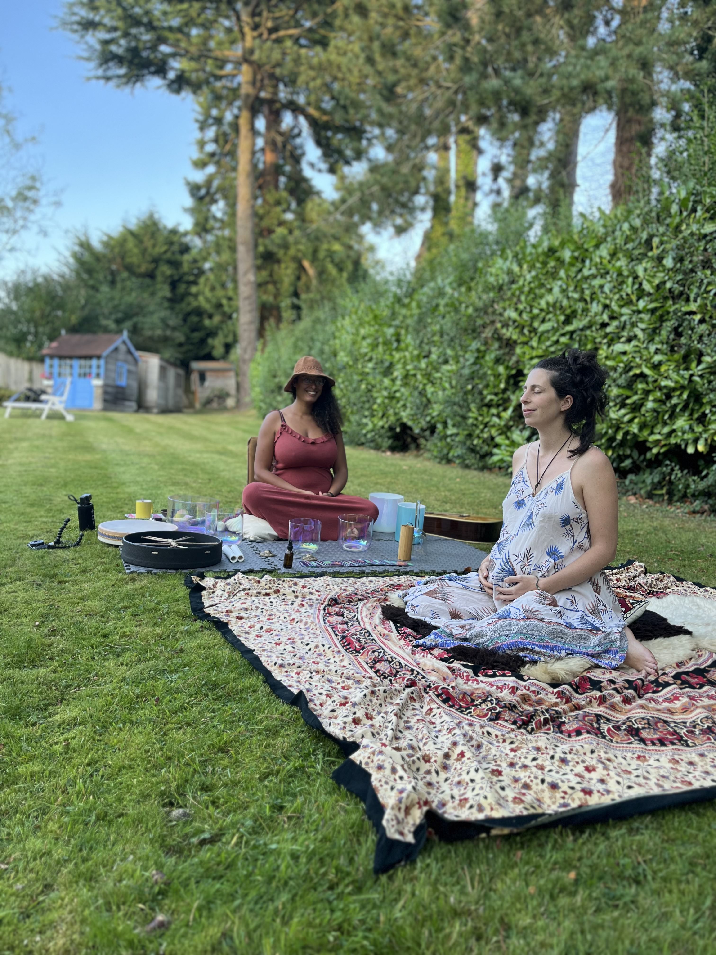 Two women sitting in a garden on green grass with a green bush behind them. The woman in the front is pregant and sits holding her belly about to have a sound bath