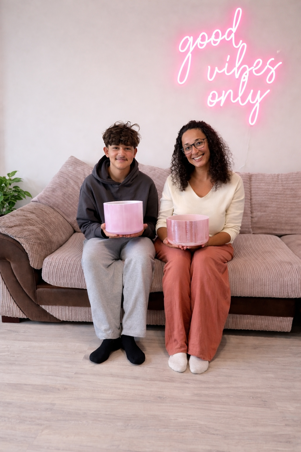 Teenage boy and Casey sitting on the sofa each holding a pink cystal tones bowl and smiling. A pink good vibes only sign behind them