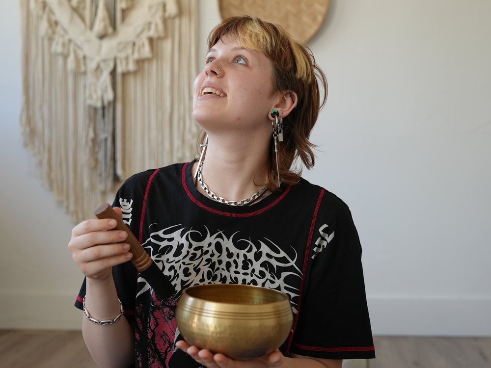 Person in a black top with red & white decorations is holding a golden tibetan singing bowl and wooden mallet. They are looking upwards and to the left. In the background is a cream macreme hanging