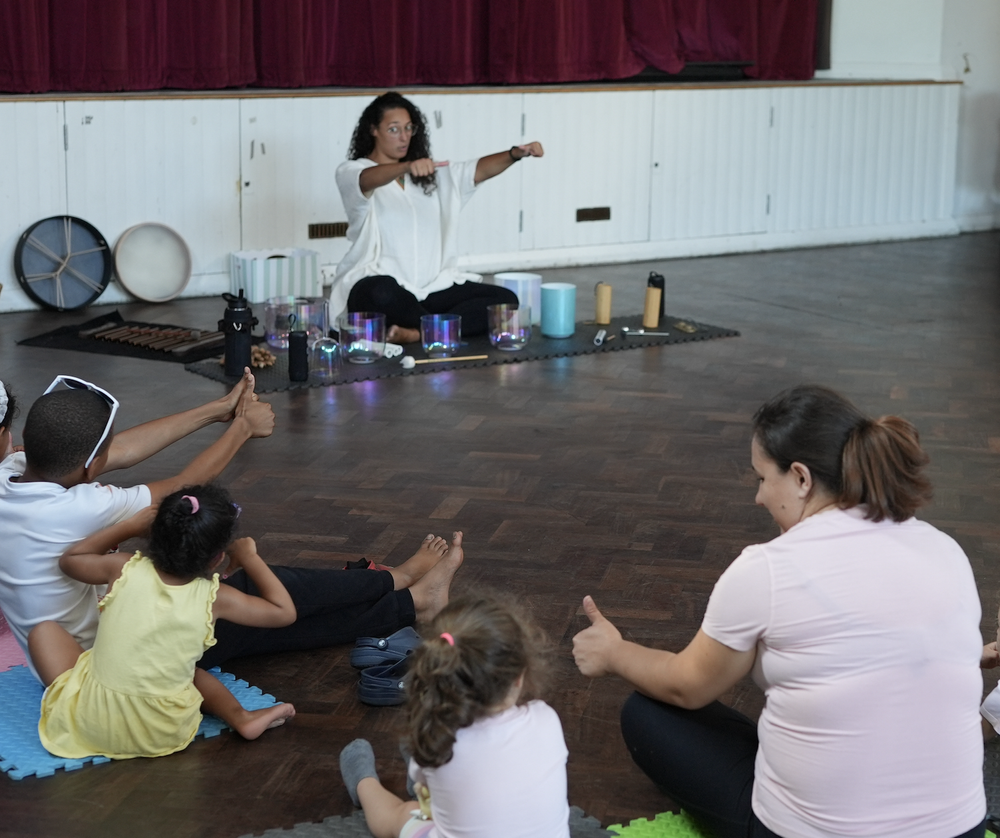 Children and a mother sitting on the floor in a sound bath giving the teacher thumbs up. The teacher wears a white top and sits behind sound healing instruments